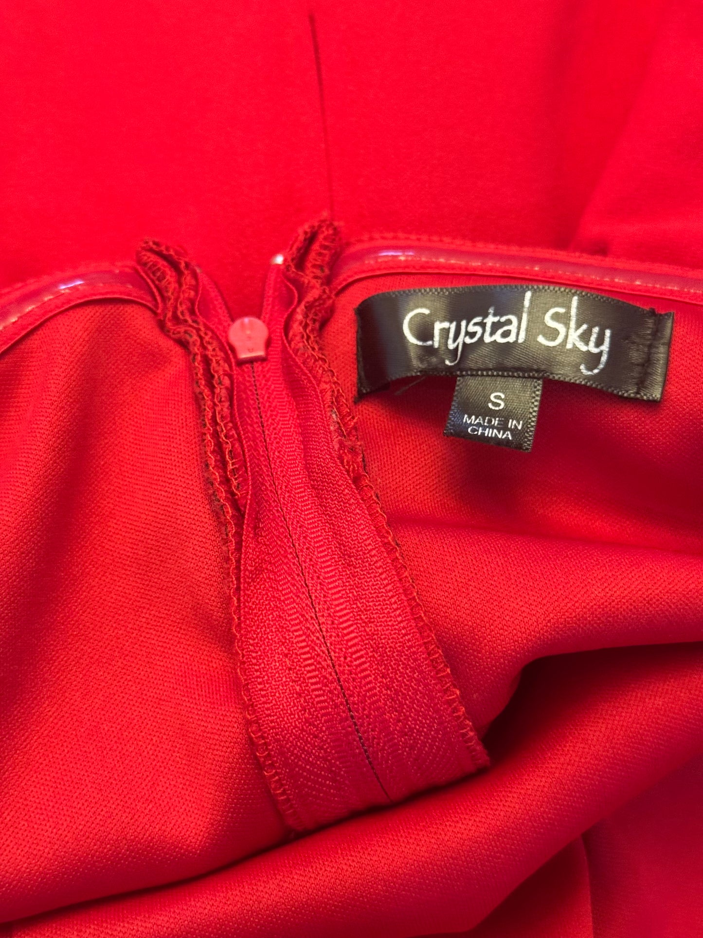 Close-up of a bright red garment with a black brand label reading Crystal Sky and a size tag reading S Made in China.