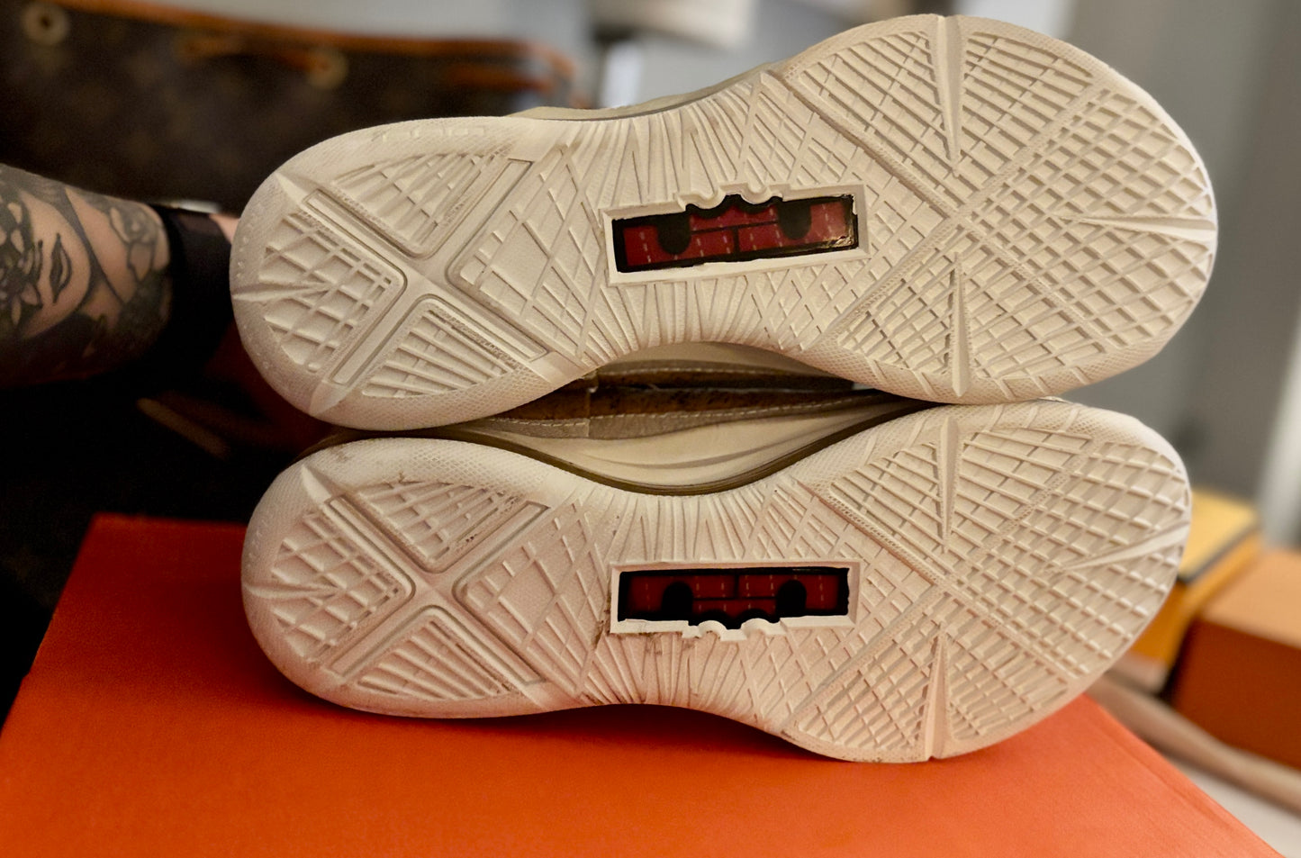 Close-up of the outsoles of a pair of beige athletic shoes, showing textured tread patterns and red translucent heel inserts, resting on an orange surface.