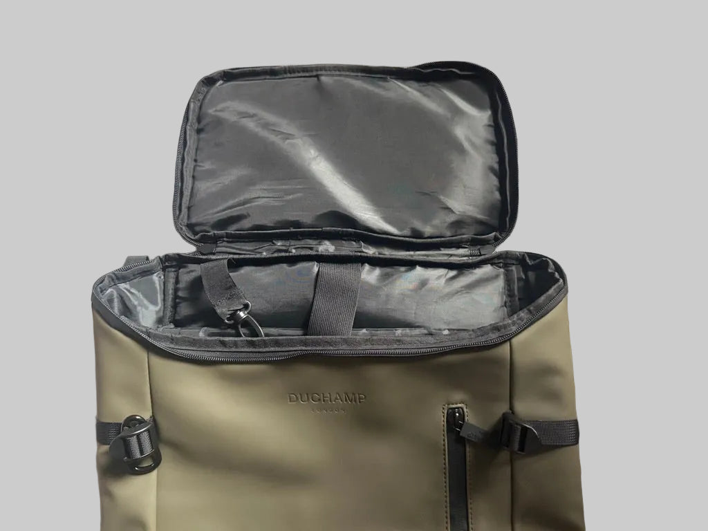 An olive green Duck Camp backpack shown open, revealing a black interior organizer pouch and zippered compartments, placed on a textured light gray blanket.