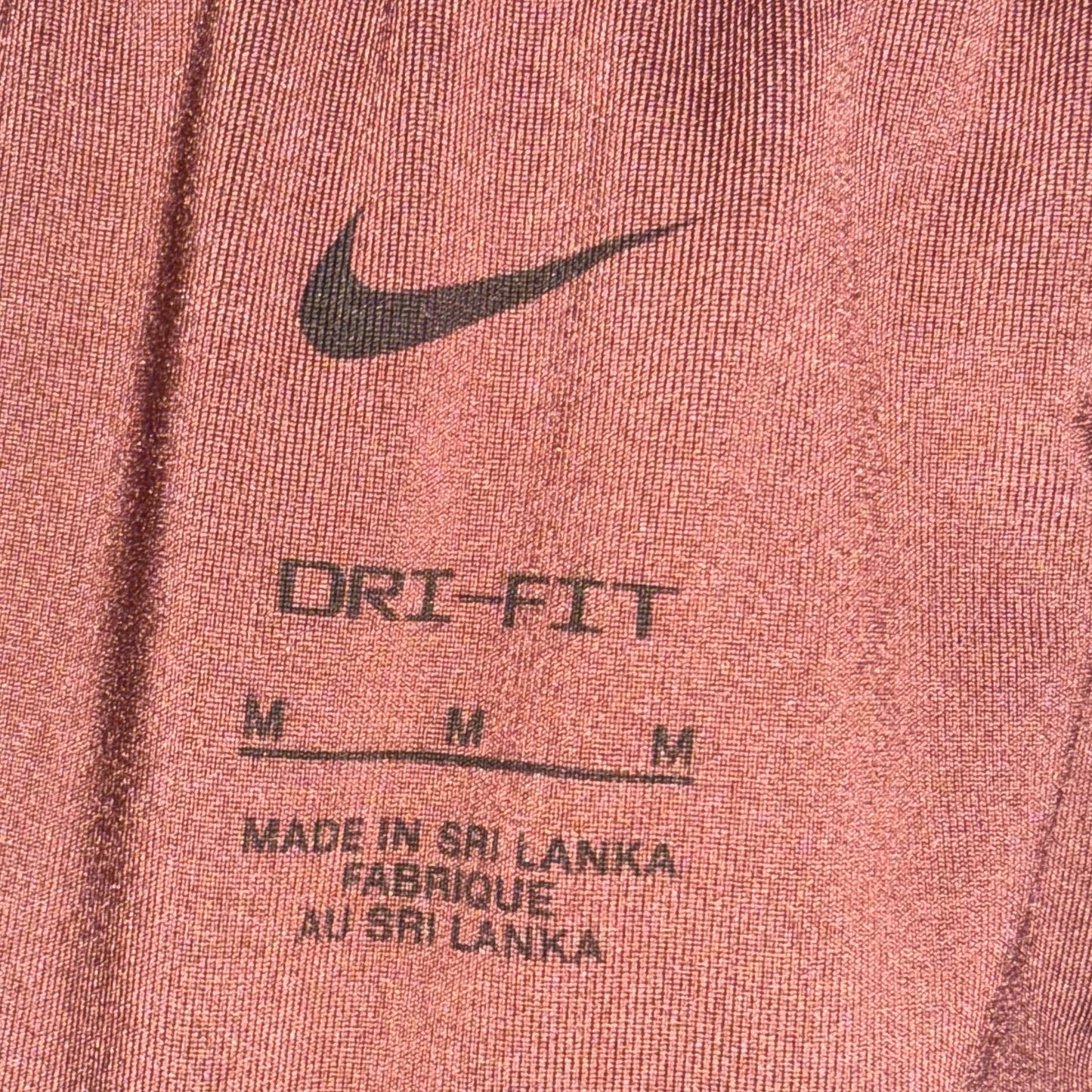 Close-up of a Nike garment, likely sweatpants or shorts, showing the elastic waistband, a black Nike swoosh, and printed text reading 'ORI-FIT' with size markings and 'Made in Sri Lanka' on a heathered dusty rose fabric.