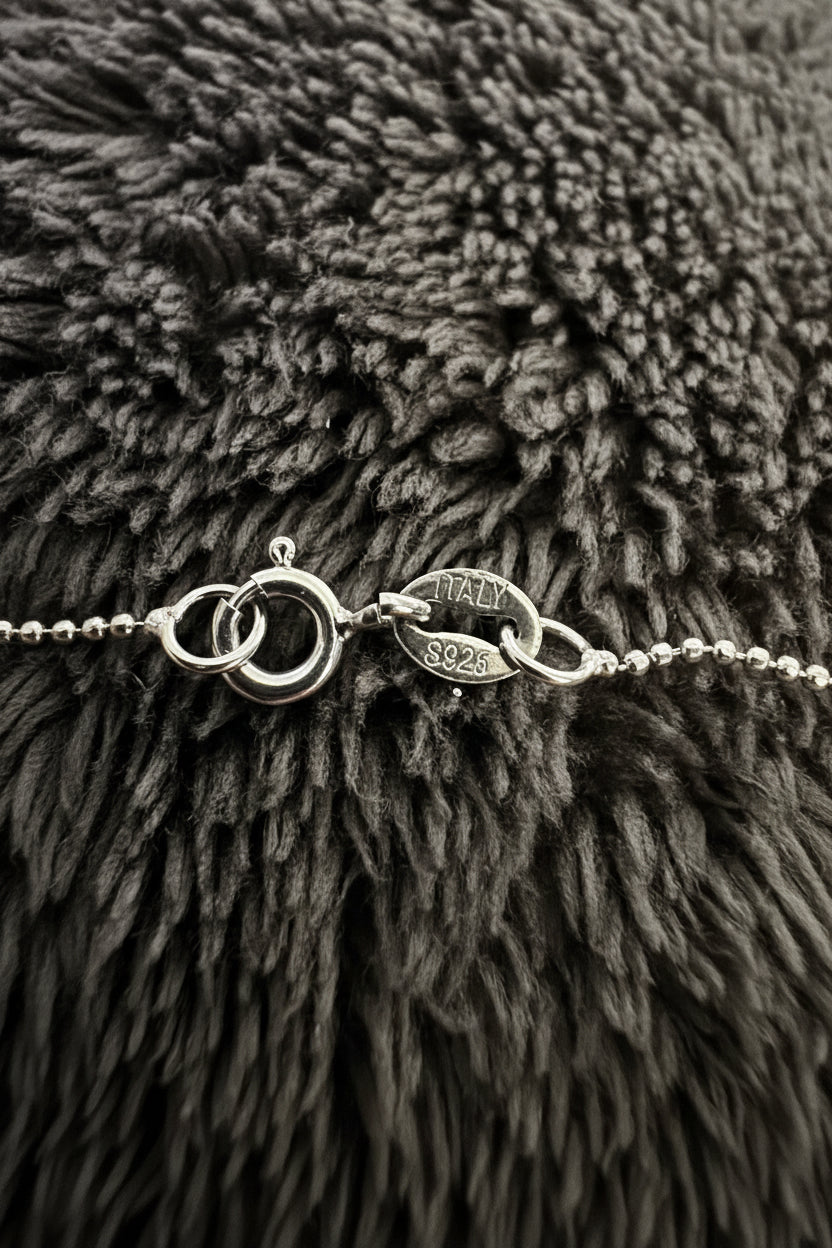 Close-up of a silver-toned chain bracelet with a lobster clasp and oval link detail, resting on a dark gray shaggy textured surface. The oval link is engraved with 'ITALY 925'.