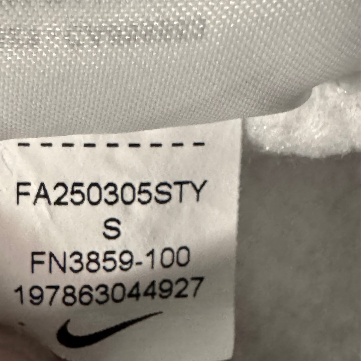 Close-up of a white garment tag with black printed text and a Nike swoosh logo, held by a finger against a gray fabric background.