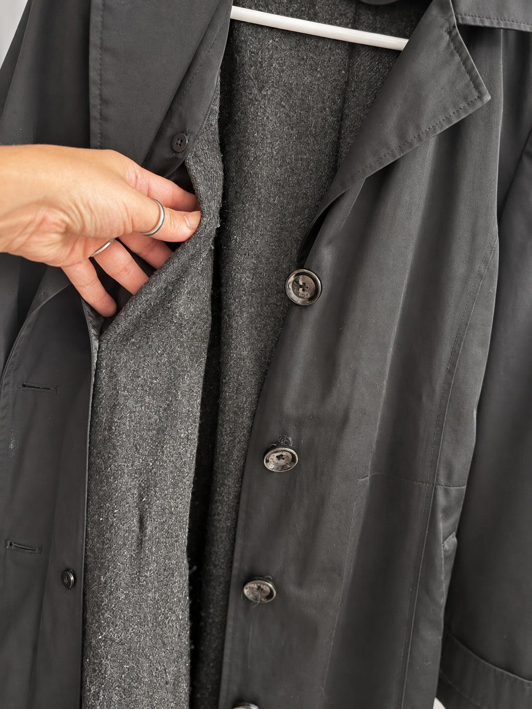 Close-up of a dark charcoal gray coat with a hand holding open the front to reveal a textured gray wool-like lining, shown on a hanger.