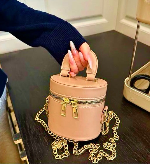 Beige handbag with gold chain held by a hand on a dark surface.