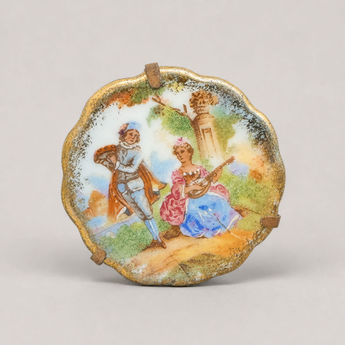 A scalloped-edge decorative plate or dish featuring a hand-painted classical scene of a man and woman in an outdoor landscape, displayed on a neutral light gray background.