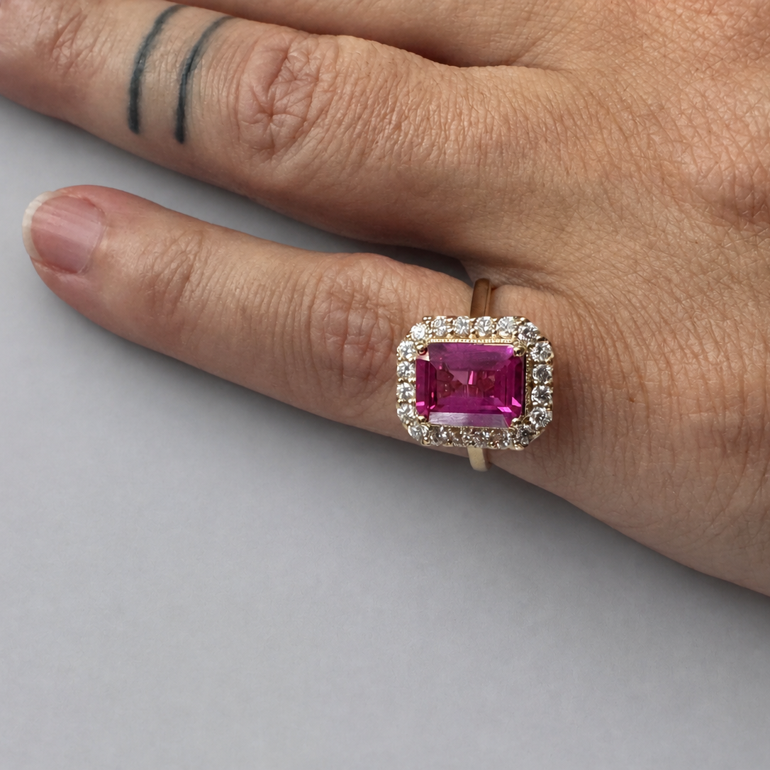A close-up of a hand wearing a yellow gold ring with a rectangular pink gemstone surrounded by a halo of small clear stones, set against a plain light gray background.