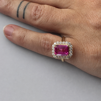 A close-up of a hand wearing a yellow gold ring with a rectangular pink gemstone surrounded by a halo of small clear stones, set against a plain light gray background.