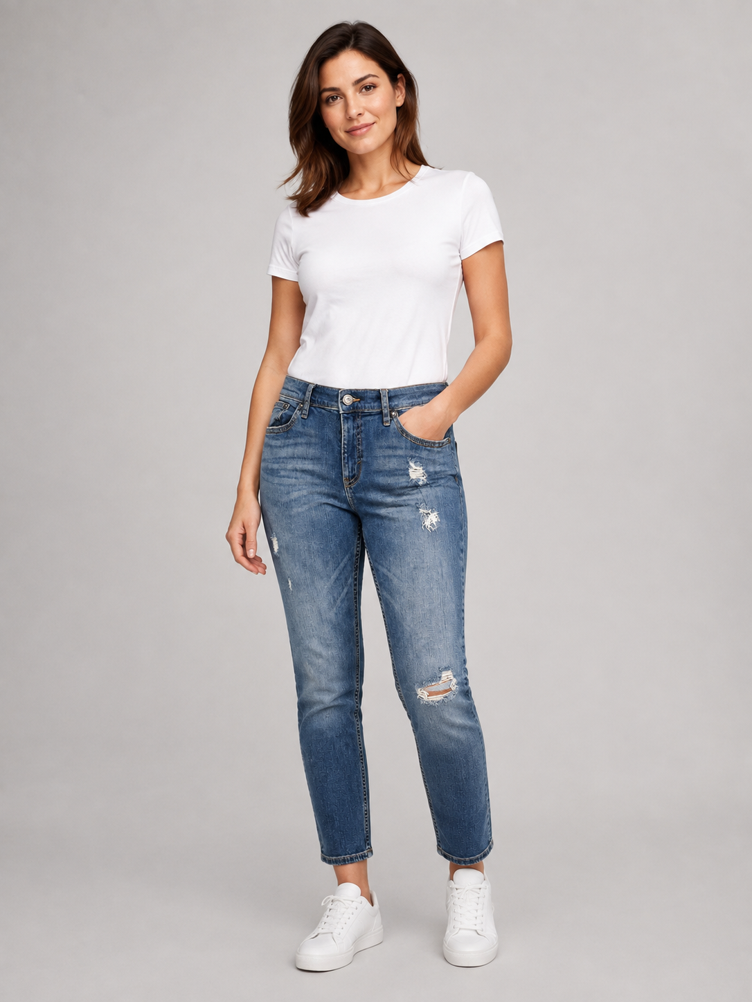 A woman modeling a pair of blue distressed skinny jeans with a white t-shirt and white sneakers, standing against a solid gray studio background.