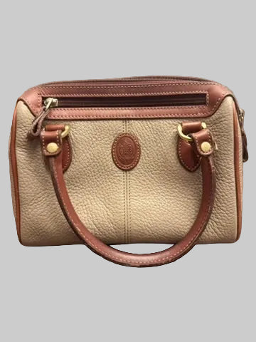 A small pebbled leather handbag in beige and brown, featuring dual top handles, a top zip closure, and a circular embossed logo patch on the front, resting on a dark wood floor.
