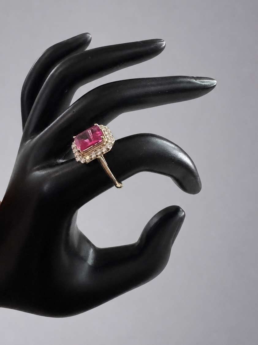 A yellow gold ring with a rectangular pink gemstone surrounded by small clear stones, displayed on a black hand-shaped stand against a neutral gray background.
