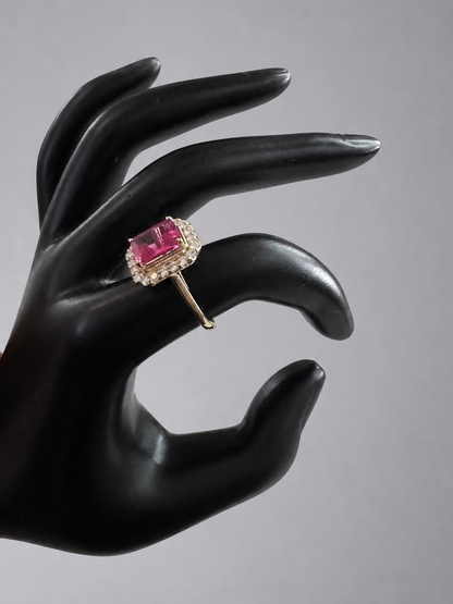 A yellow gold ring with a rectangular pink gemstone surrounded by small clear stones, displayed on a black hand-shaped stand against a neutral gray background.