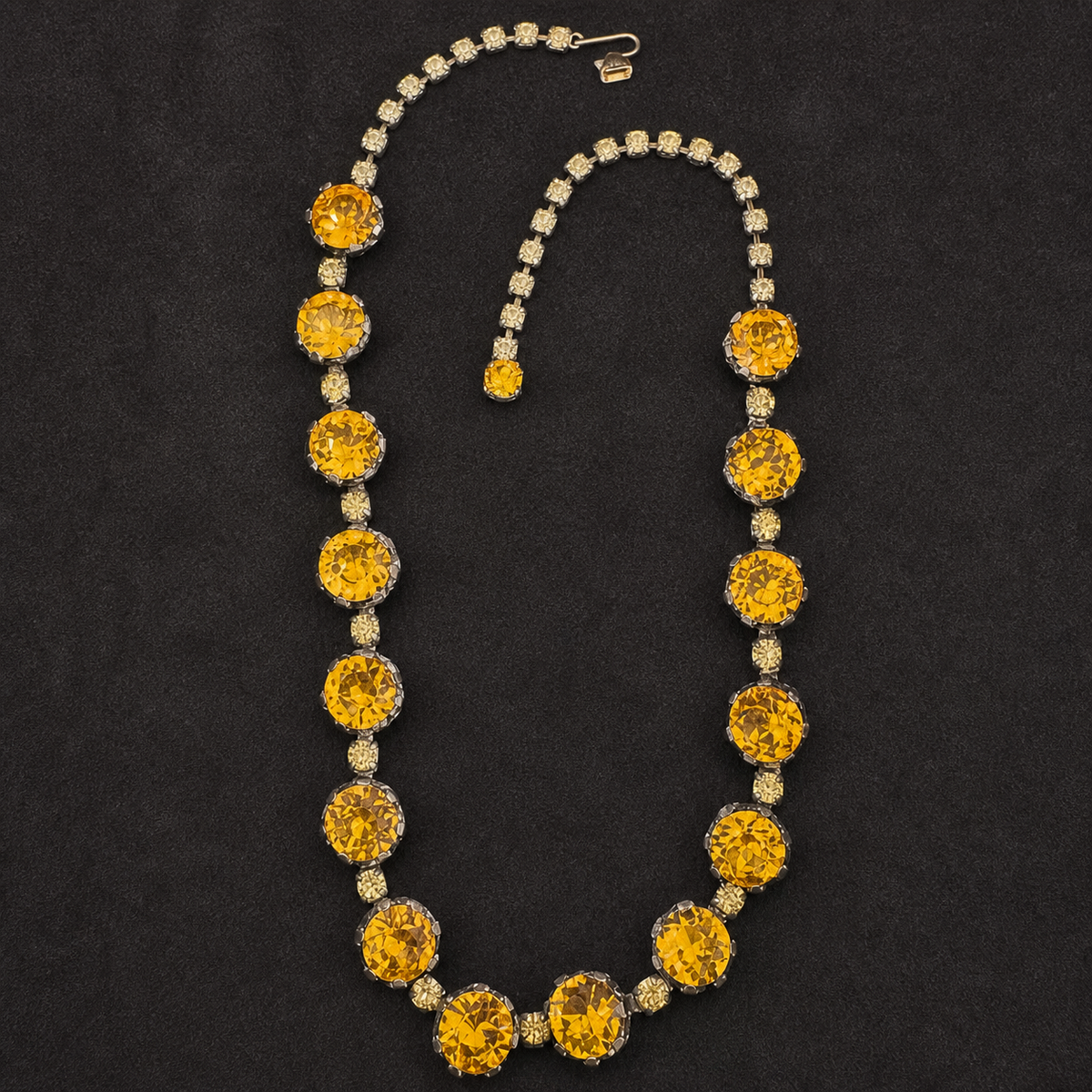 A vintage-style necklace with large round yellow gemstones and smaller clear stones, displayed on a dark textured background. A matching small pendant is placed beside the necklace.