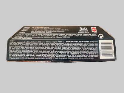 The back of a Barbie-branded product box, showing printed text, a barcode, and a small red Barbie logo, photographed against a blurred indoor background.