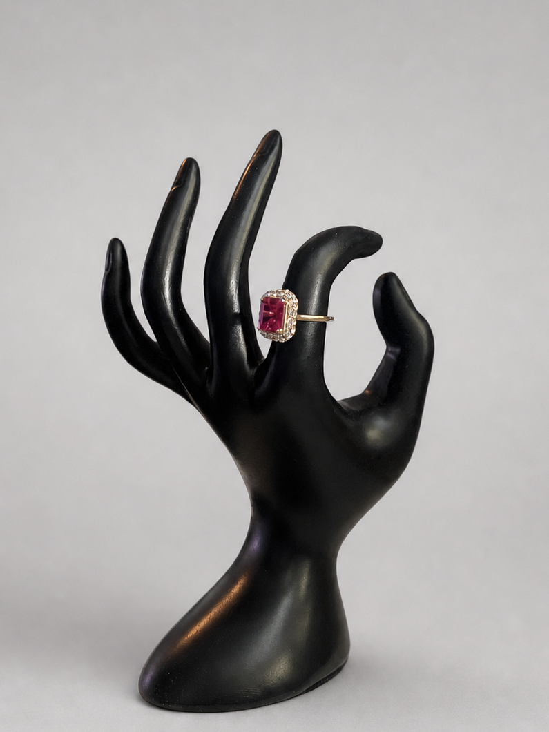A gold ring with a round pinkish-red gemstone displayed on a black hand-shaped jewelry stand against a light gray background.