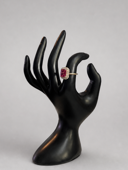 A gold ring with a round pinkish-red gemstone displayed on a black hand-shaped jewelry stand against a light gray background.