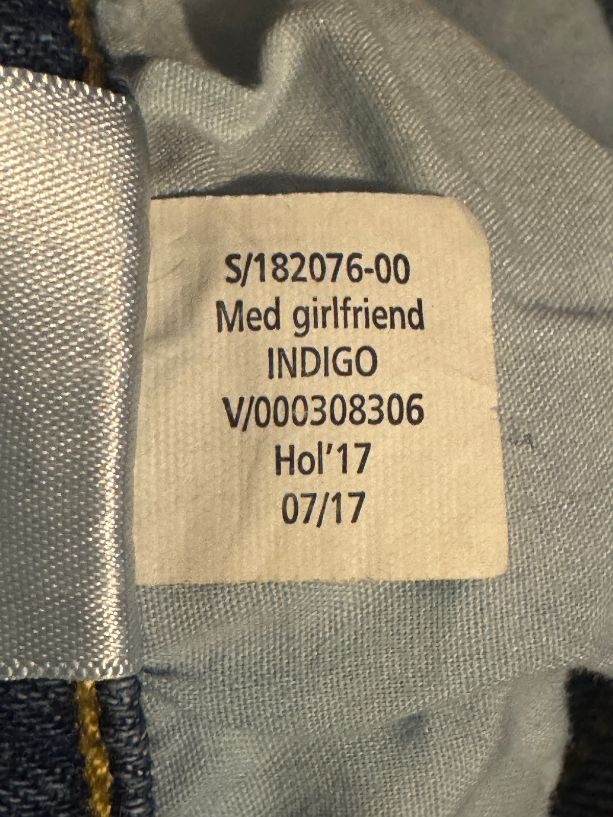 Close-up of a sewn-on size and care label inside a pair of jeans, showing printed text including 'S/182076-00 Med girlfriend INDIGO V/000308306 Holi'17 07/17' on light gray fabric.