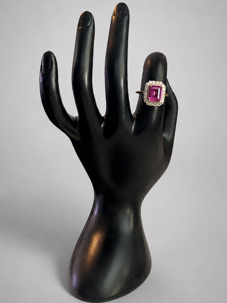 A ring with a rectangular pink gemstone in a silver-toned setting, displayed on a black hand-shaped jewelry stand against a light gray background.