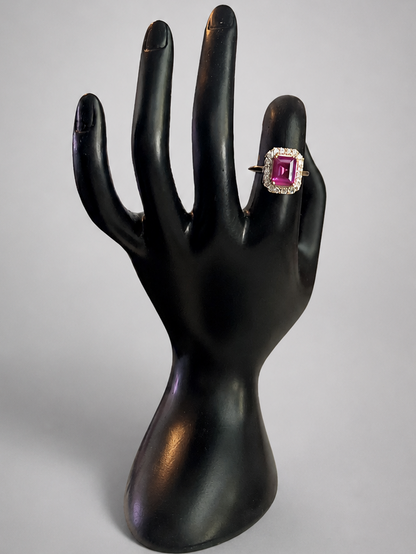 A ring with a rectangular pink gemstone in a silver-toned setting, displayed on a black hand-shaped jewelry stand against a light gray background.