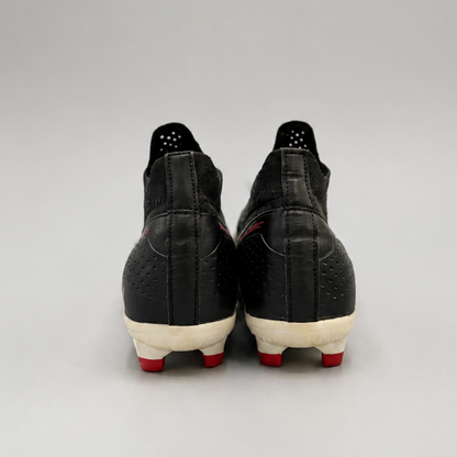 A pair of black soccer cleats shown from the back, featuring textured black uppers, off-white midsoles, and red outsole spikes, set against a neutral gray studio background.