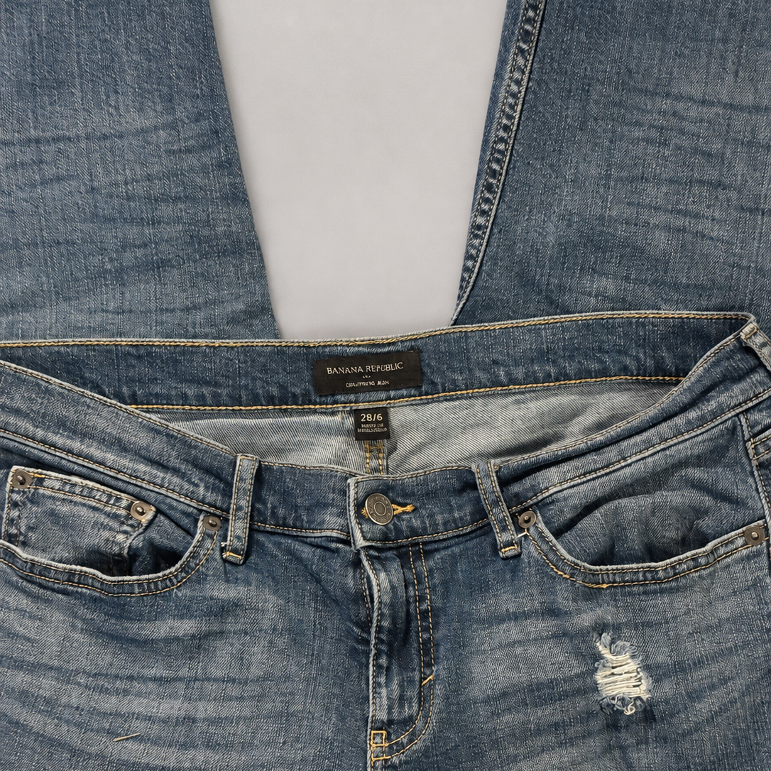 Close-up of folded blue denim jeans showing the waistband, belt loops, front pockets, button, rivets, and a small distressed rip on the left leg, photographed on a light gray background.