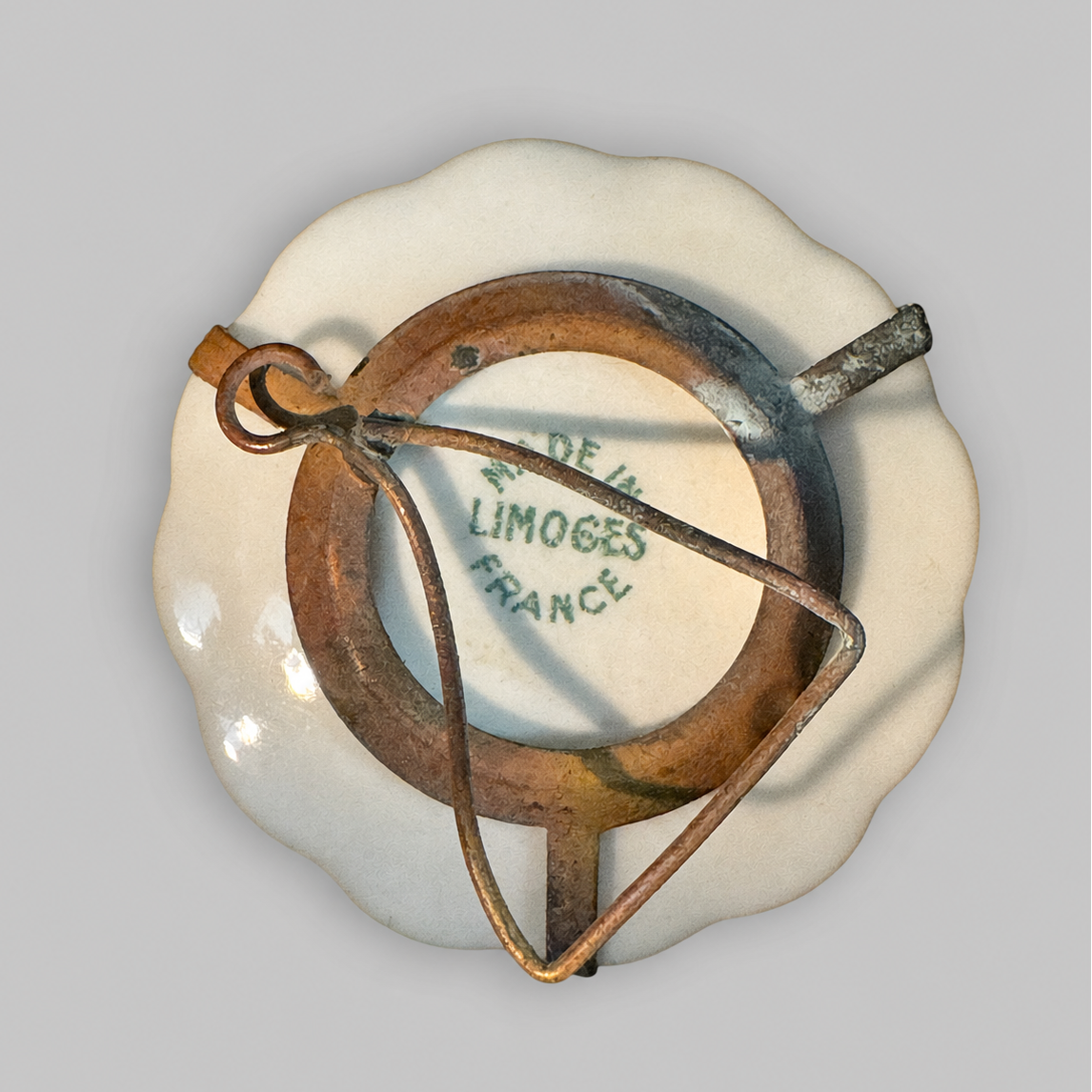 An antique French Limoges porcelain pin badge with a scalloped cream-colored border, a central brown ring, and a rusted metal loop, showing the 'DE LA LIMOGES FRANCE' mark on the inside.