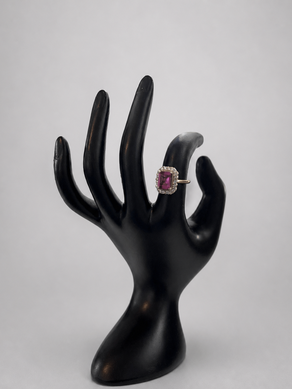 A ring with a rectangular pink gemstone and a silver-toned setting, displayed on a black hand-shaped jewelry stand against a light gray background.