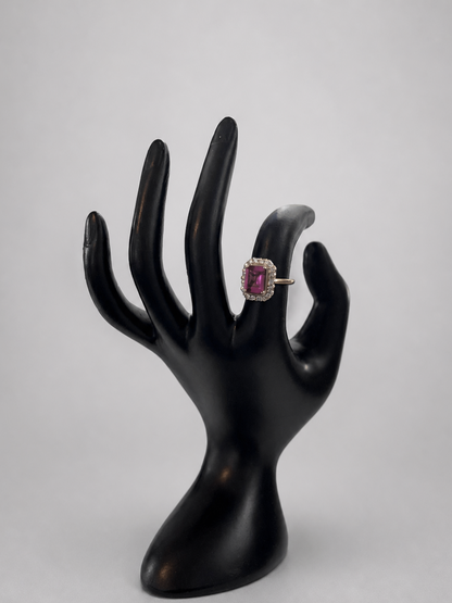 A ring with a rectangular pink gemstone and a silver-toned setting, displayed on a black hand-shaped jewelry stand against a light gray background.