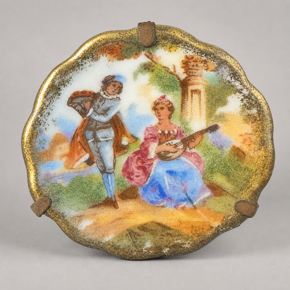 A scalloped antique ceramic plate or decorative dish featuring a hand-painted classical scene of a man carrying a bundle and a woman playing a lute, surrounded by a distressed gold rim on a neutral gray background.