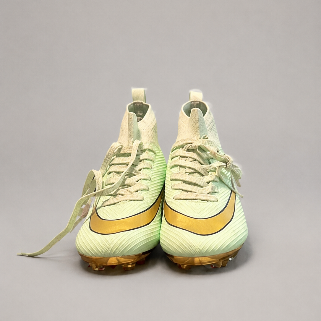 A pair of  high-top soccer cleats in pale mint green and gold, shown from the front on a neutral gray background.
