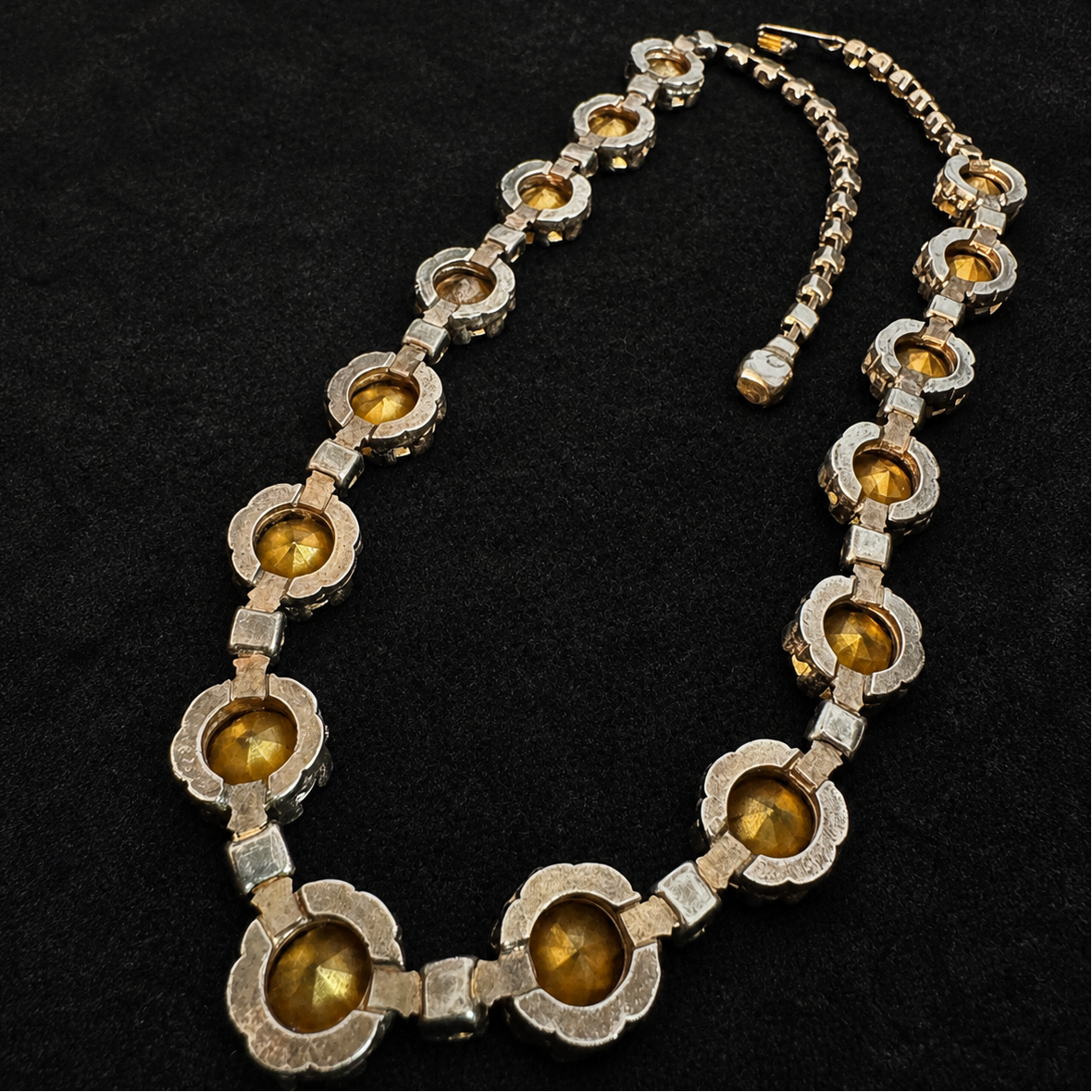 A vintage-style statement necklace featuring scalloped silver-tone metal links with round amber-yellow faceted gemstones, displayed on a black textured background.