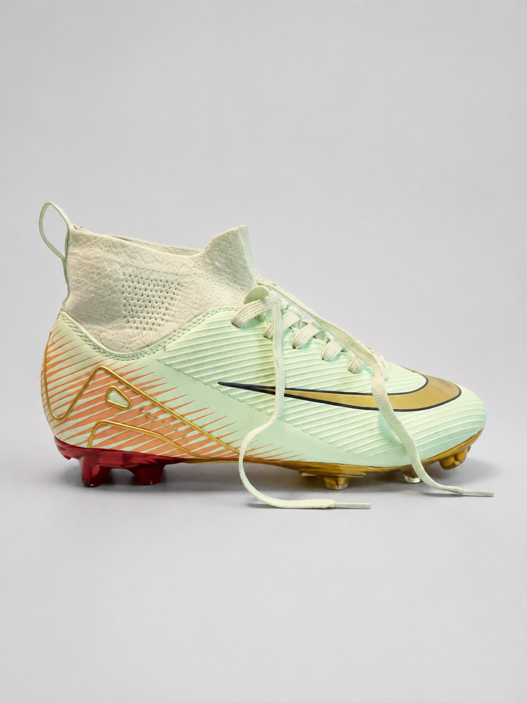 A soccer cleat in a pale mint green and gold colorway, featuring a knit sock-like collar, orange and gold diagonal accents on the heel, and red and gold studs.