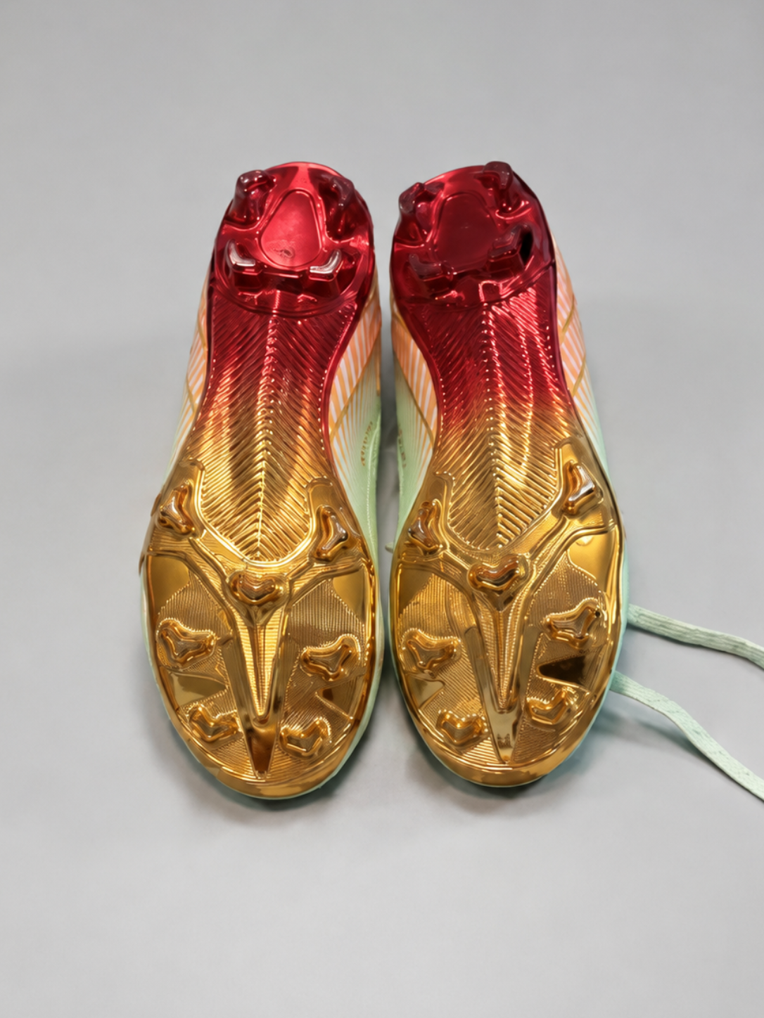 A pair of soccer cleats with a metallic gold and red gradient upper, featuring a light mint green sole and white laces, photographed on a neutral gray background.