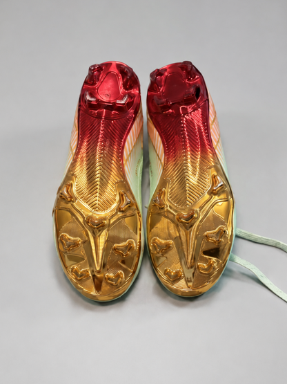 A pair of soccer cleats with a metallic gold and red gradient upper, featuring a light mint green sole and white laces, photographed on a neutral gray background.