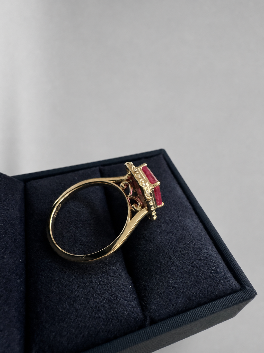 A gold ring with a rectangular red gemstone and decorative filigree setting, displayed inside an open dark navy velvet ring box on a light gray background.