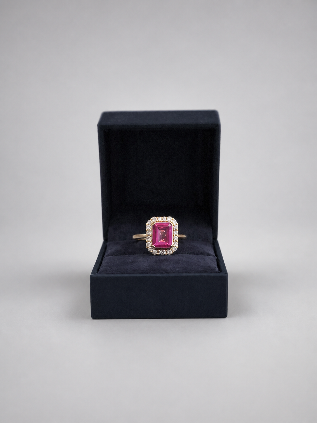 A rectangular pink gemstone ring with a gold halo and band, displayed inside an open black velvet ring box on a neutral gray background.