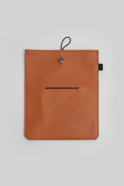 An orange-brown leather tablet sleeve or document holder with a black hanging loop and a horizontal slit, laid flat on a gray textured rug.