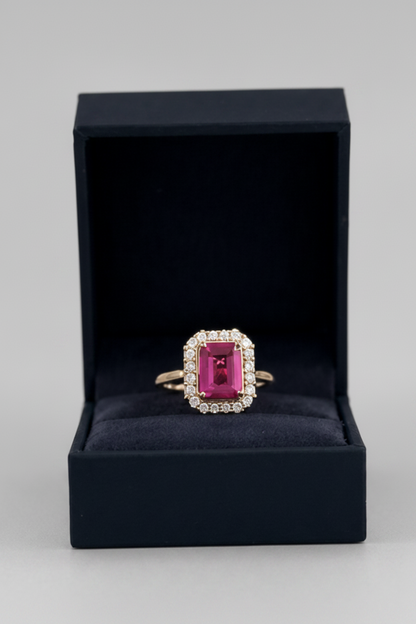 A yellow gold ring with a rectangular emerald-cut pink gemstone and a halo of small clear stones, displayed in an open black jewelry box on a light gray background.
