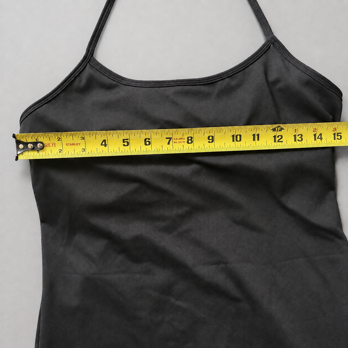 A black camisole tank top shown against a light gray background with a yellow tape measure stretched horizontally across the underbust area to indicate the garment's waist or bust measurement.
