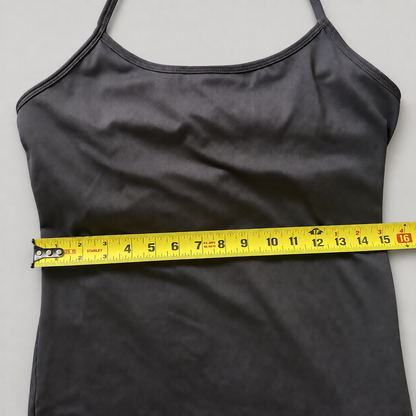 A dark gray camisole tank top laid flat on a light gray background, with a yellow tape measure stretched horizontally across the waist area to show the garment's circumference.