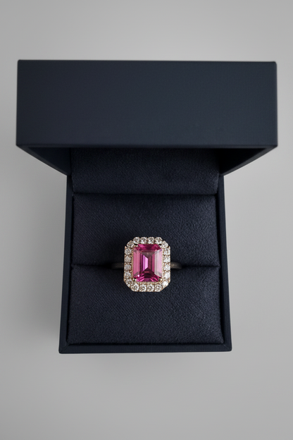 A rectangular emerald-cut pink gemstone ring with a halo of small clear stones, displayed in an open dark velvet jewelry box against a neutral gray background.