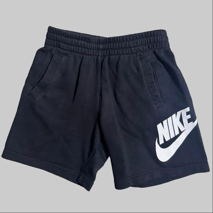 Black Nike athletic shorts with a white logo printed on the left leg, featuring an elastic waistband and side pockets, shown against a solid gray background.