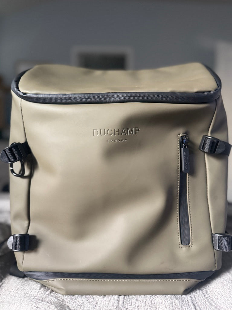 A beige waterproof backpack by Duchamp London with black trim, side straps, and a vertical zipper pocket, resting on a textured white surface.