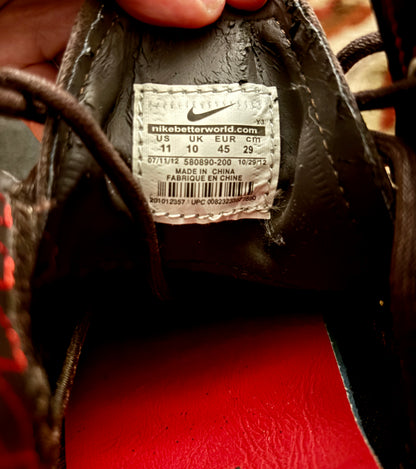 Close-up of the inside of a Nike shoe showing the size and model label, with a hand holding the shoe. The interior is dark brown, with a red footbed visible at the bottom.