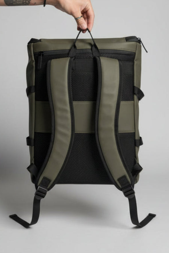 An olive green and black backpack shown from the back, held by a hand in a bedroom setting with a bed and TV visible in the background.