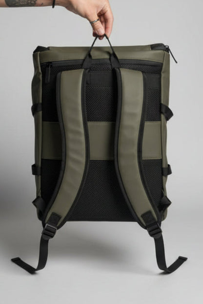 An olive green and black backpack shown from the back, held by a hand in a bedroom setting with a bed and TV visible in the background.
