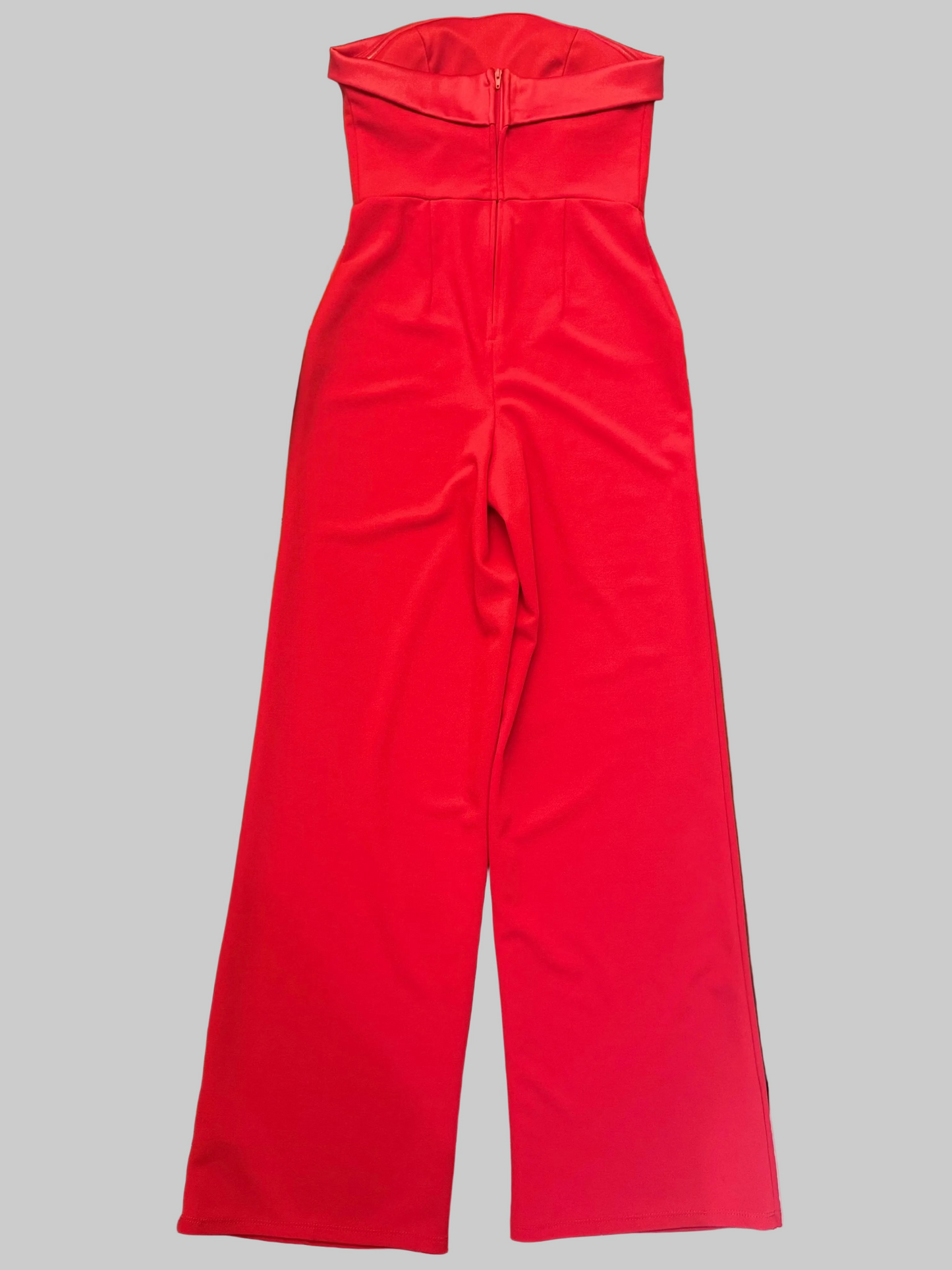 A bright red strapless jumpsuit with a wide waistband and wide legs, laid flat on a wooden table with various household items around it.