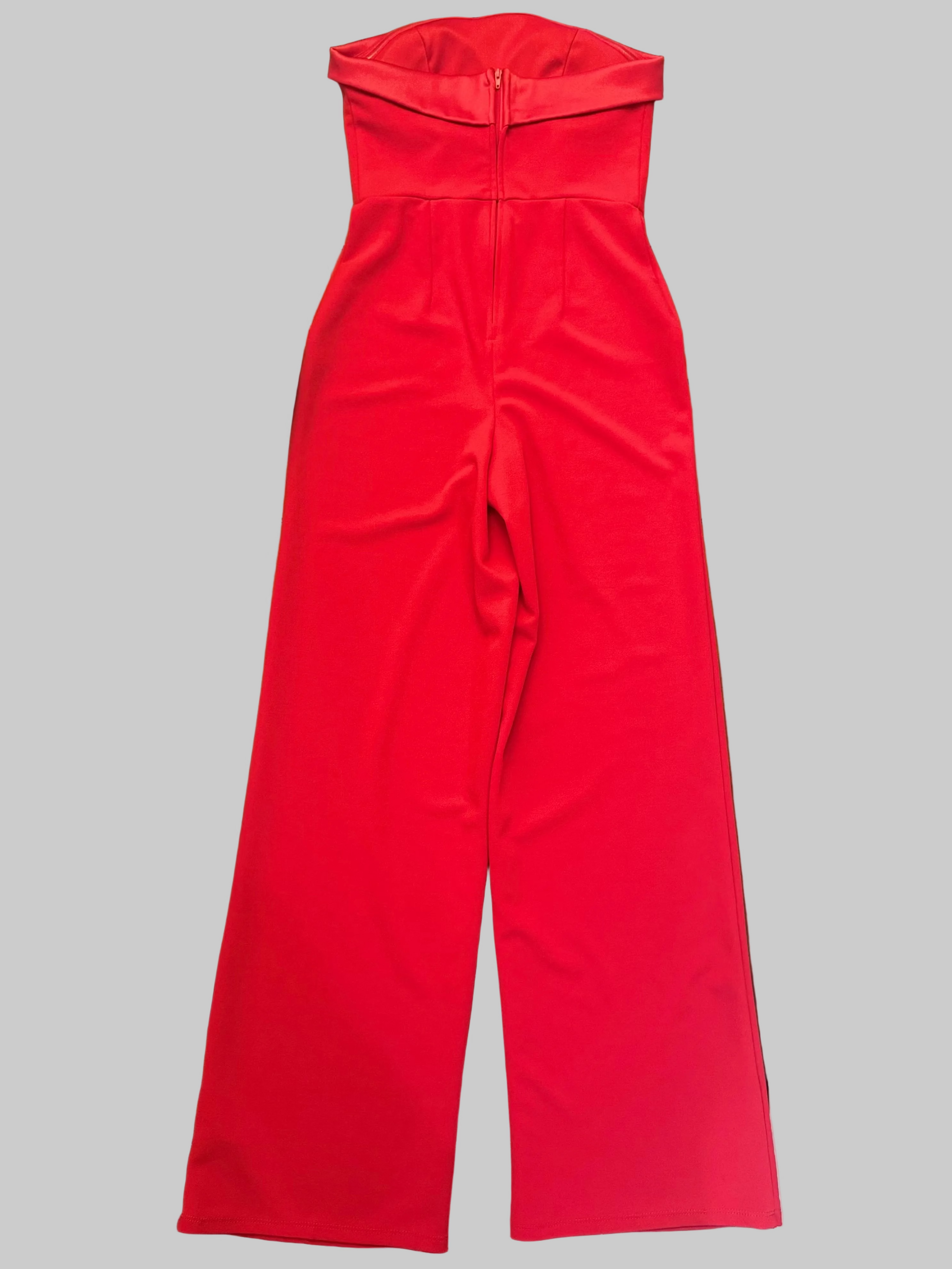 A bright red strapless jumpsuit with a wide waistband and wide legs, laid flat on a wooden table with various household items around it.