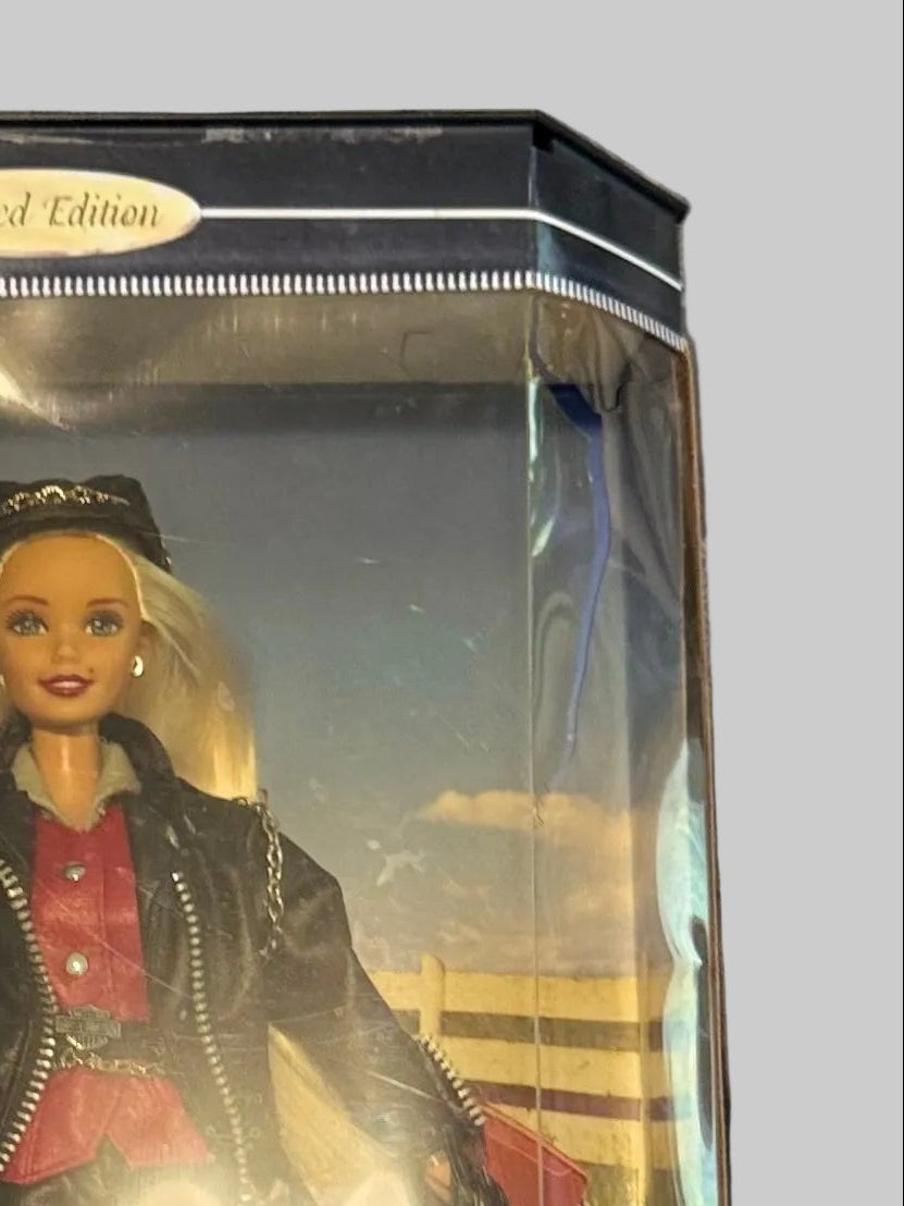 A boxed Barbie doll dressed as a motorcycle rider, standing in front of a Harley-Davidson logo on the packaging. The package is photographed on a plaid blanket.