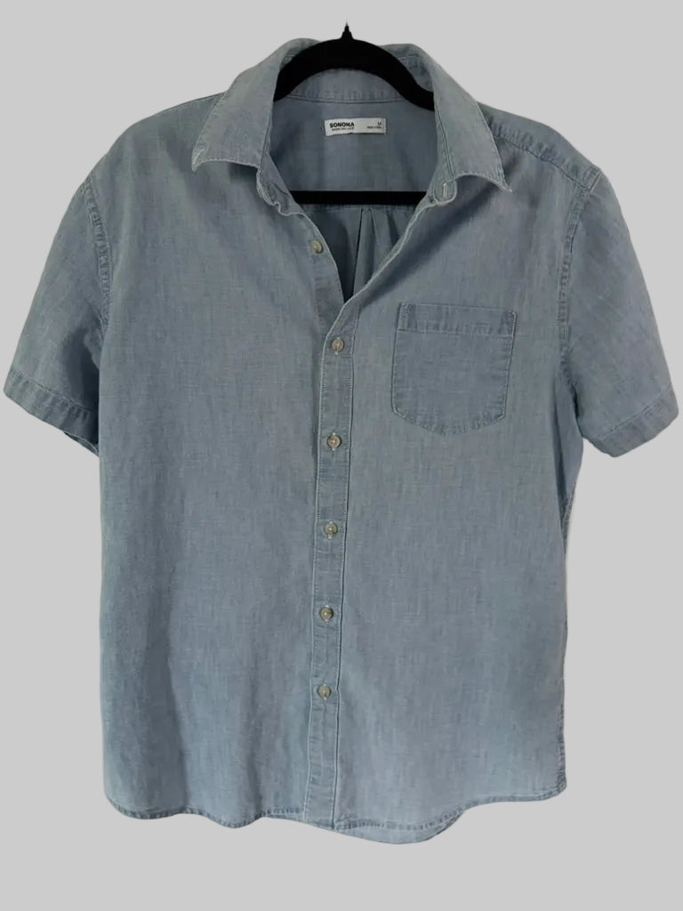 A light blue chambray short-sleeve button-up shirt hanging on a black hanger against a white paneled door.