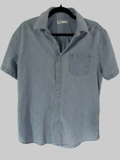 A light blue chambray short-sleeve button-up shirt hanging on a black hanger against a white paneled door.
